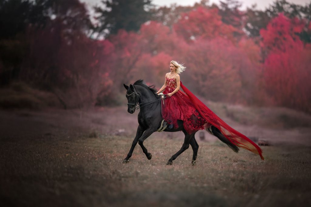 Eine Frau in rotem Kleid reitet auf einem schwarzen Pferd durch eine herbstliche Landschaft.
