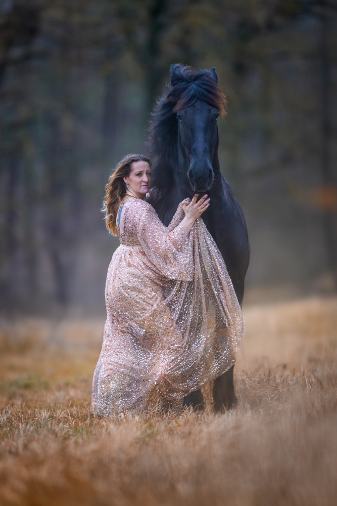 Frau in sanftem Kleid umarmt ein schwarzes Pferd in einer herbstlichen Landschaft.