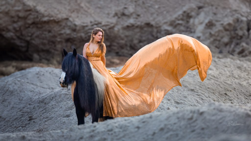 Frau in orangenem Kleid steht neben einem schwarzen Pferd in einer sandigen Landschaft.