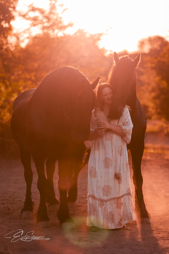 Frau in einem hellen Kleid, umgeben von zwei großen Pferden bei Sonnenuntergang.