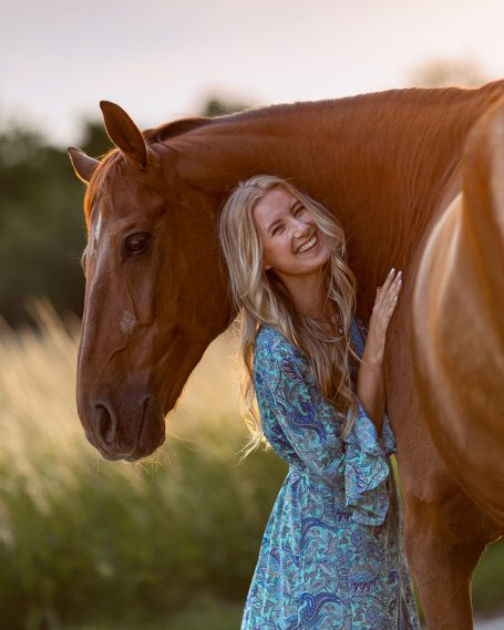 Junge Frau mit blonden Haaren umarmt ein braunes Pferd in einer sonnigen Wiesenlandschaft.