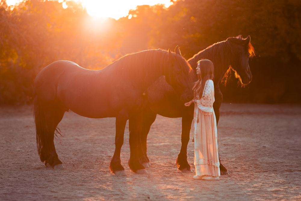 Frau in weißem Kleid neben zwei großen, dunklen Pferden bei Sonnenuntergang.